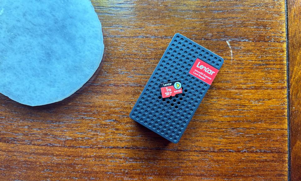 A red microSD Express card sits on top of a small black microSD card reader, on top of a brown wooden table, next to a white stone drink coaster.